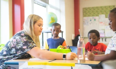 Teacher writes at a table with three young students in a colorful classroom.