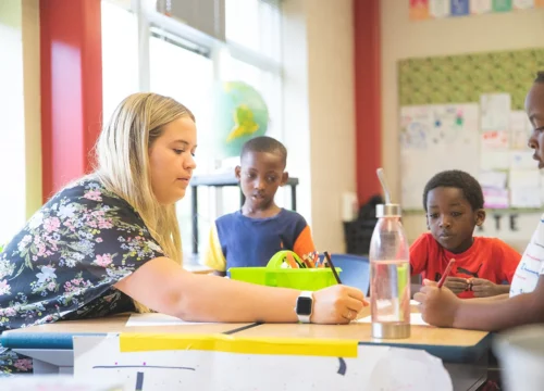 Teacher writes at a table with three young students in a colorful classroom.