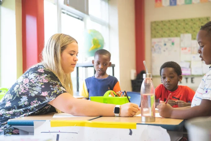 Teacher writes at a table with three young students in a colorful classroom.
