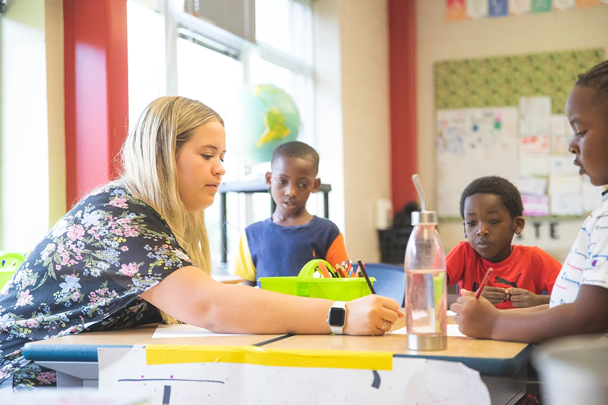 Teacher writes at a table with three young students in a colorful classroom.
