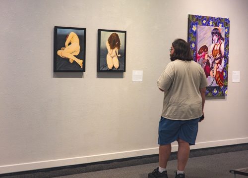 A person stands looking at two pieces of art with painted human figures on a wall. A third piece of art is on the wall to the right.