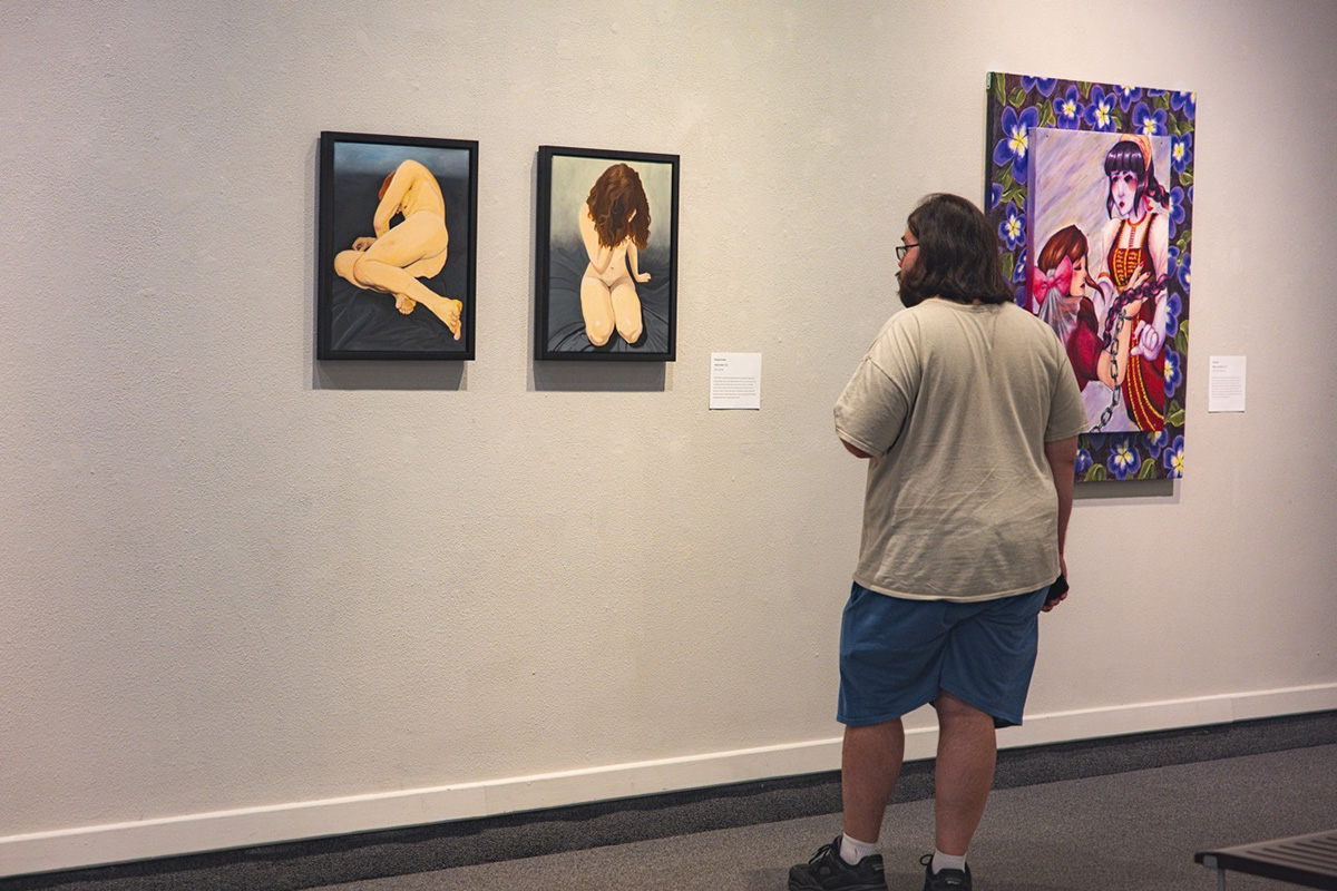 A person stands looking at two pieces of art with painted human figures on a wall. A third piece of art is on the wall to the right.
