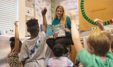 Teacher sits on a stool while reading a book to a group of elementary students sitting on the floor. Two students have their hands raised.