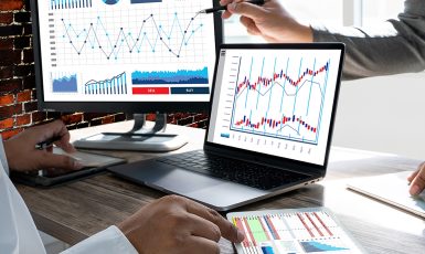 Charts and graphs on a computer monitor, laptop and printed paper with two hands pointing at them.