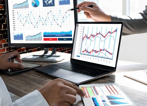 Charts and graphs on a computer monitor, laptop and printed paper with two hands pointing at them.