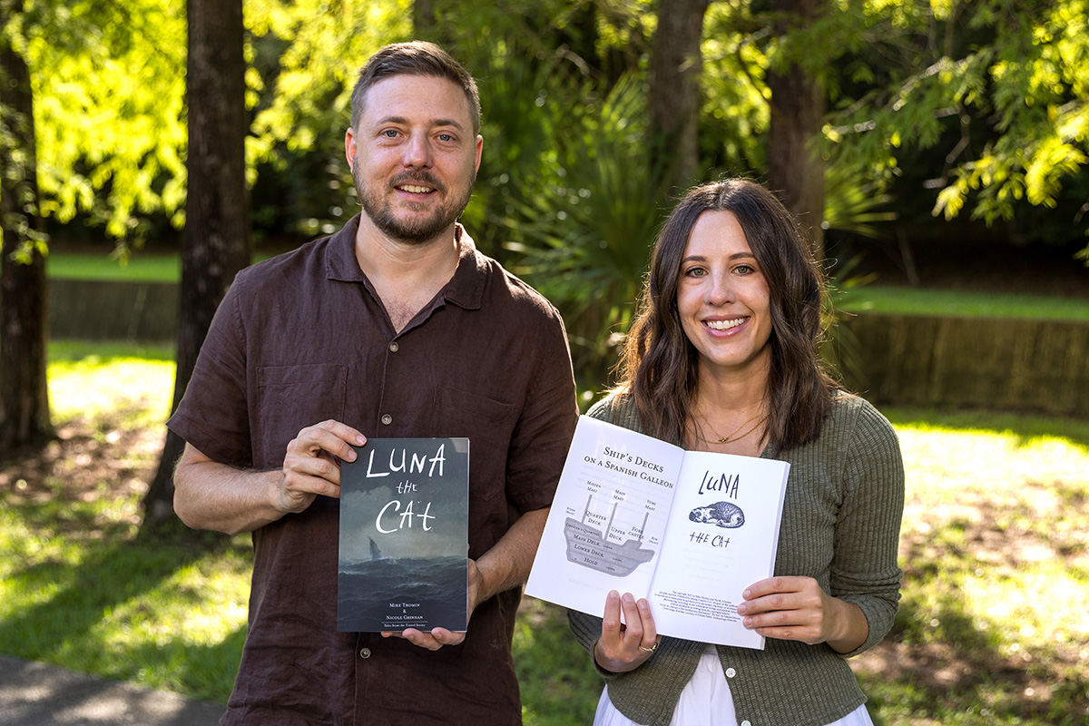 Two people stand side by side smiling. One holds a book showing the cover that says, "Luna the Cat" and the other holds a book open to pages.