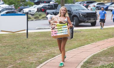 A smiling person holds a box while walking down a path from the parking lot.