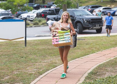 A smiling person holds a box while walking down a path from the parking lot.