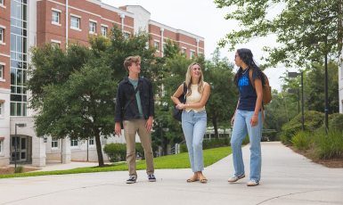 Three students walk along a sidewalk on the UWF campus in front of a building.