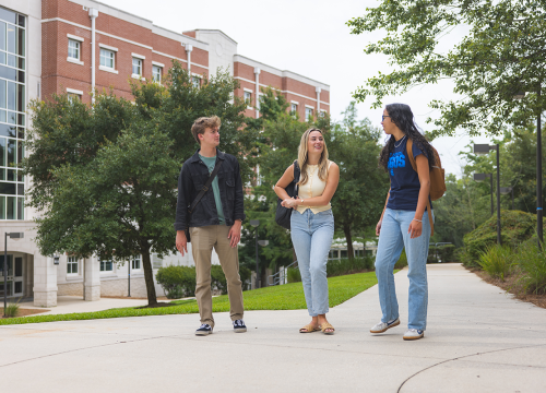 Three students walk along a sidewalk on the UWF campus in front of a building.