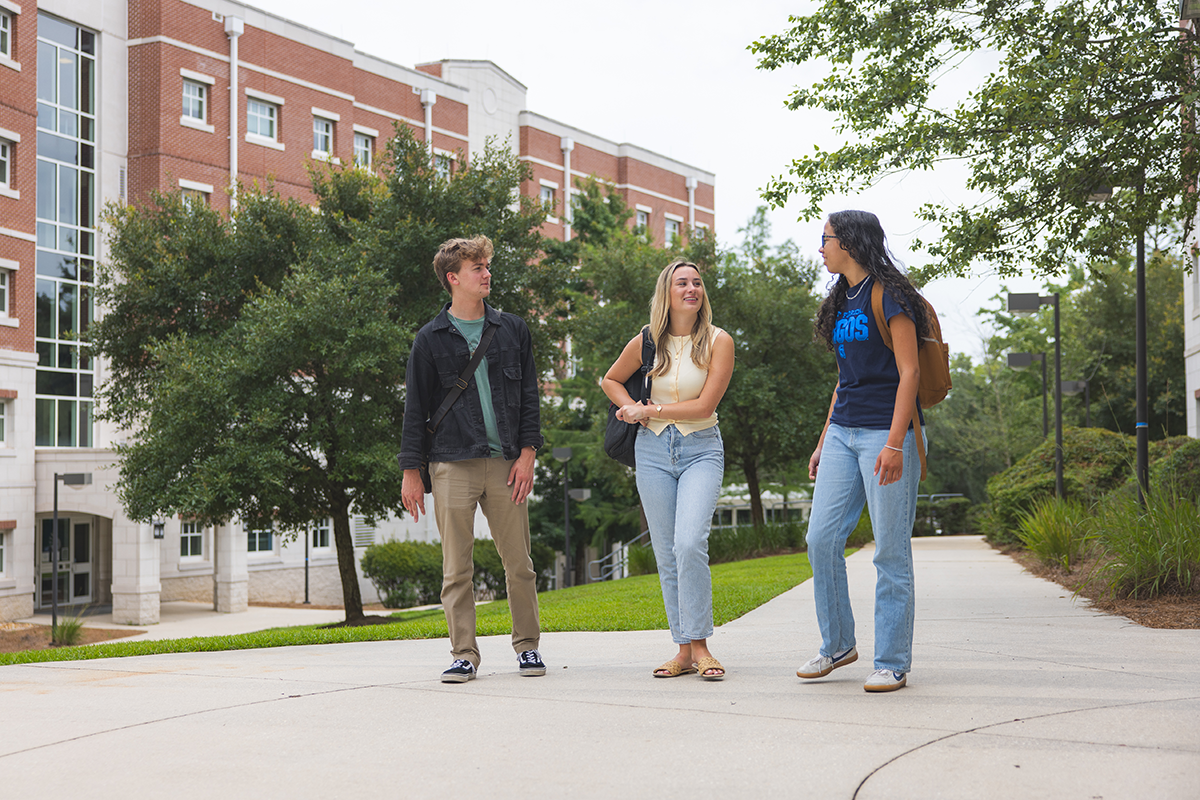 Three students walk along a sidewalk on the UWF campus in front of a building.