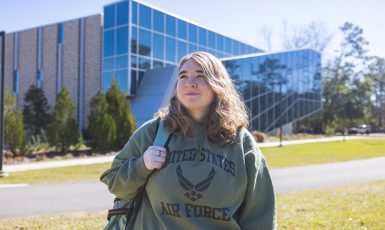 Student in United States Air Force hoodie smiles off into the distance while holding a backpack strap with one arm.