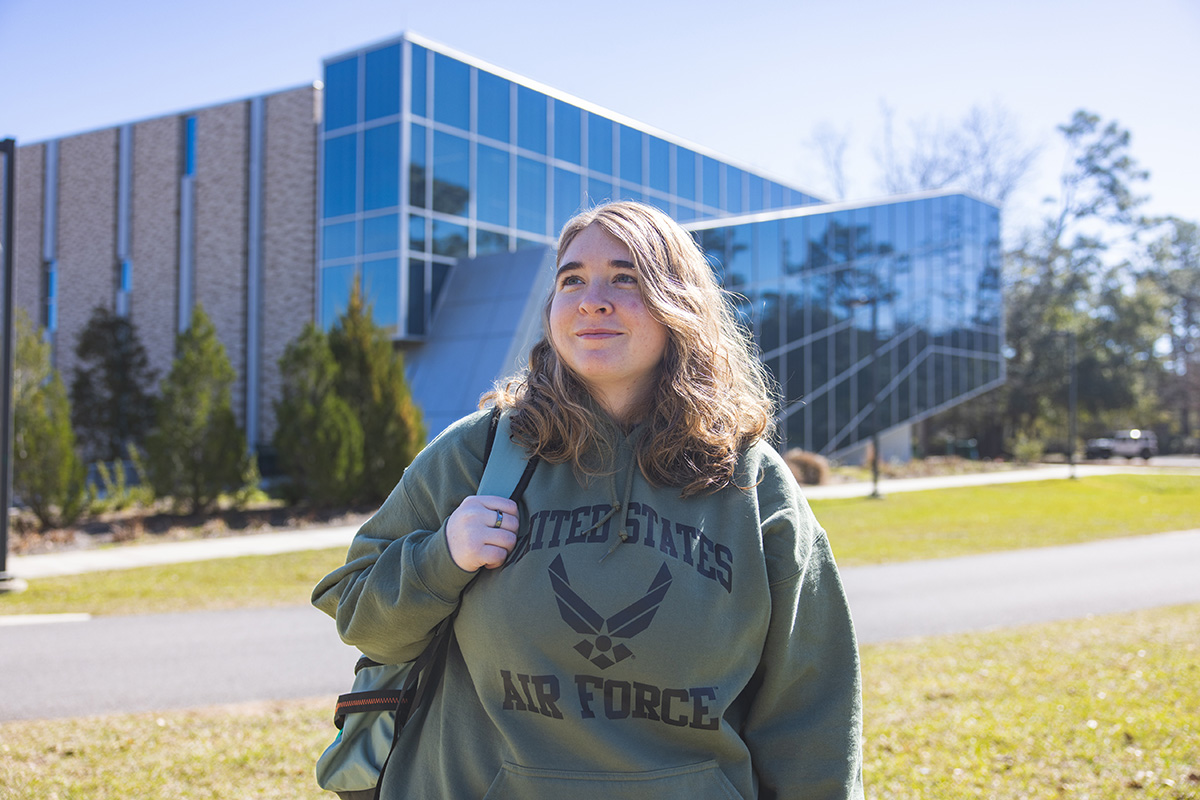Student in United States Air Force hoodie smiles off into the distance while holding a backpack strap with one arm.