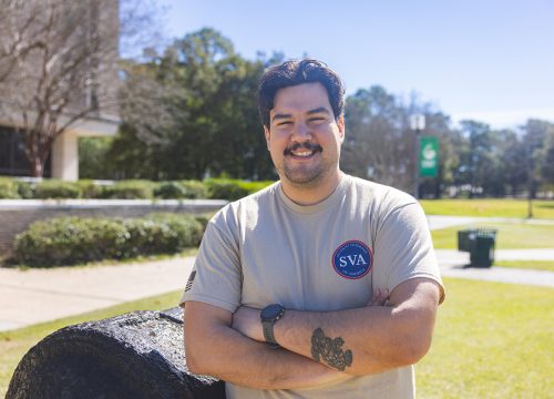 UWF student veteran stands with arms crossed smiling in front of the cannon on the UWF Cannon Green