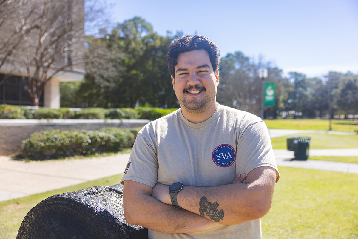 UWF student veteran stands with arms crossed smiling in front of the cannon on the UWF Cannon Green