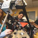 Three University of West Florida students sitting at desk working on mechanical robot arm.