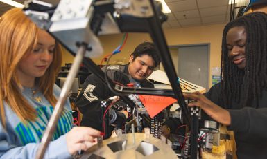 Three University of West Florida students sitting at desk working on mechanical robot arm.