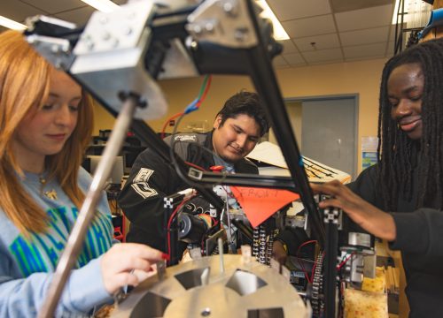 Three University of West Florida students sitting at desk working on mechanical robot arm.