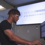 Student looks at a computer screen. A tv screen on the wall behind the person says, "Center for Cybersecurity at the University of West Florida."