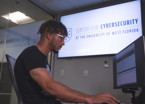 Student looks at a computer screen. A tv screen on the wall behind the person says, "Center for Cybersecurity at the University of West Florida."