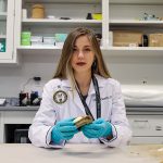 Intern in a white lab coat with a patch that reads "Defense POW/MIA Accounting Agency" in a scientific lab setting.