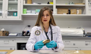 Intern in a white lab coat with a patch that reads "Defense POW/MIA Accounting Agency" in a scientific lab setting.