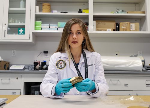 Intern in a white lab coat with a patch that reads "Defense POW/MIA Accounting Agency" in a scientific lab setting.