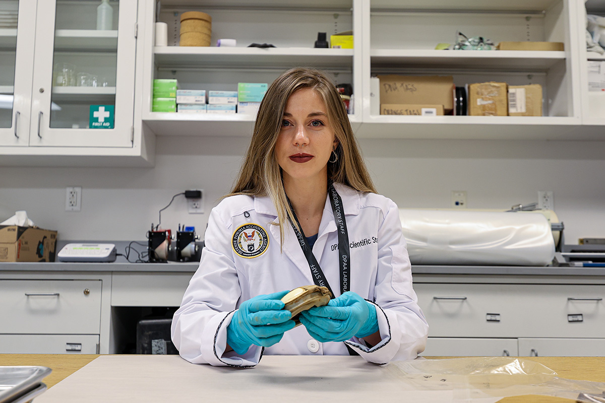 Intern in a white lab coat with a patch that reads "Defense POW/MIA Accounting Agency" in a scientific lab setting.