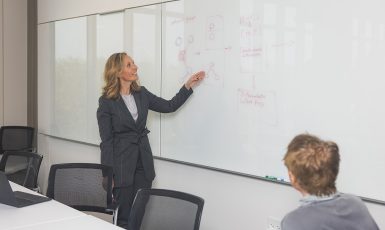 Dr. Brent Venable points to a white board with red figured drawn on it while a seated student looks.