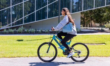 Photo: Person rides a bicycle on a sidewalk in front of a building with glass windows.