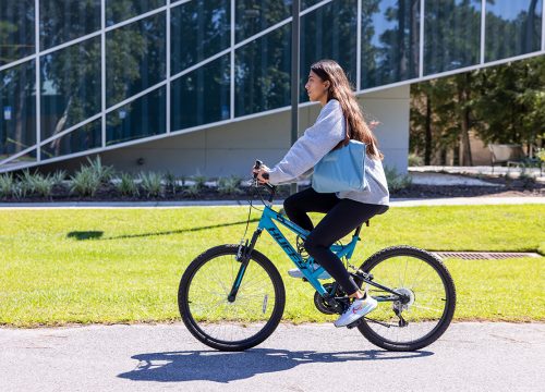 Photo: Person rides a bicycle on a sidewalk in front of a building with glass windows.