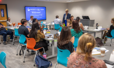 Photo: Students sit in a classroom with a smart board that reads, "University of West Florida. Welcome! Instructional Design & Performance Technology Doctoral Residency."
