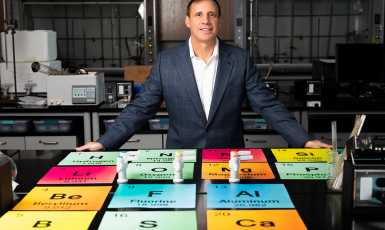 Photo: Professor stands in a lab with a giant colorful periodic table of elements displayed on a table in front of him.