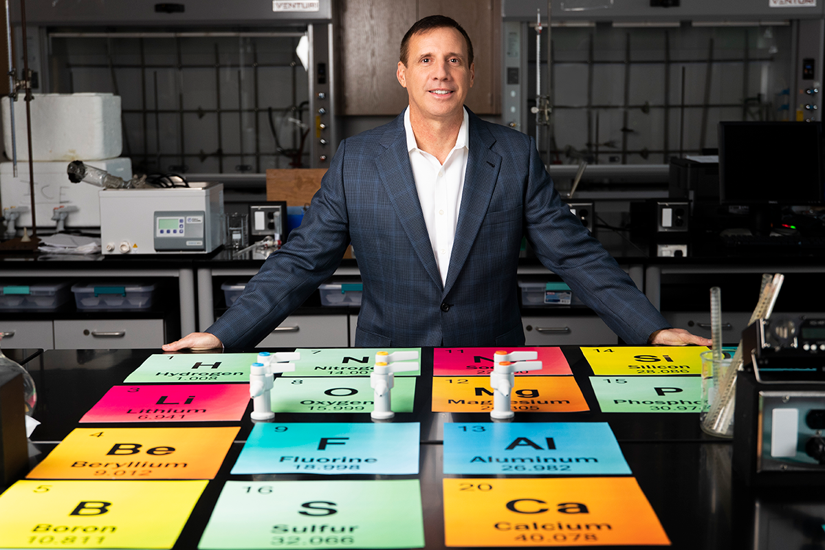 Photo: Professor stands in a lab with a giant colorful periodic table of elements displayed on a table in front of him.