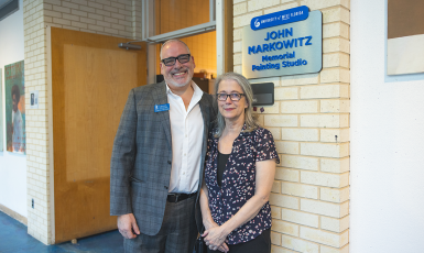 Two people pose side by side in front of a doorway and sign that reads, "University of West Florida John Markowitz Memorial Painting Studio"