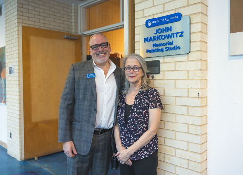 Two people pose side by side in front of a doorway and sign that reads, "University of West Florida John Markowitz Memorial Painting Studio"