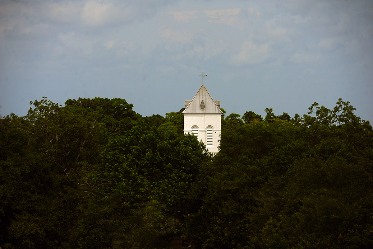 Top of a white church spire sticking out atop trees with a gray cloudy sky.