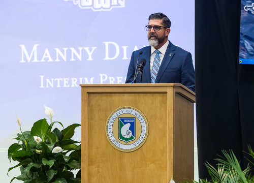 President Manny Diaz Jr. gives a speech behind a podium. Screen behind him reads, "Manny Diaz Jr. Interim President."