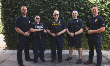 Five UWF police officers pose in front of a green shrub wall.