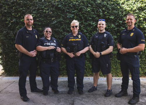 Five UWF police officers pose in front of a green shrub wall.