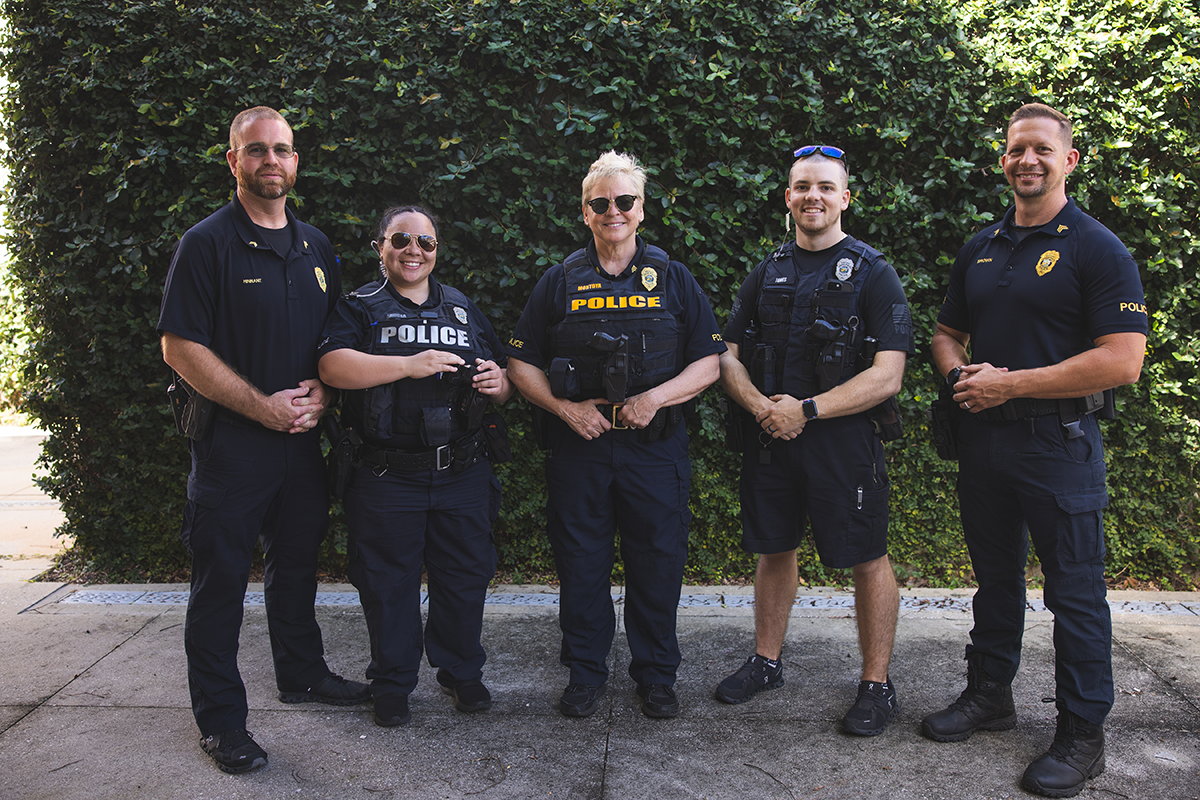 Five UWF police officers pose in front of a green shrub wall.