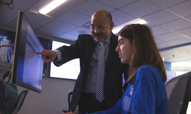 Photo: Professor points at a computer screen while a seated student looks on.