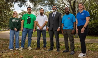 President Diaz stands beside five military affiliated students under some trees on campus.