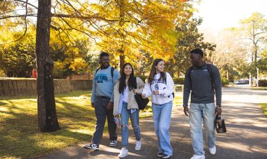 Photo: Four students walk on a sidewalk, talking and laughing, under golden trees.