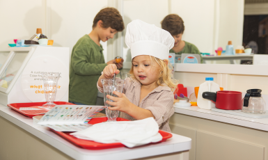 Photo: Toddler plays with milkshake glass behind a diner counter while another slightly older child plays in the kitchen behind her.