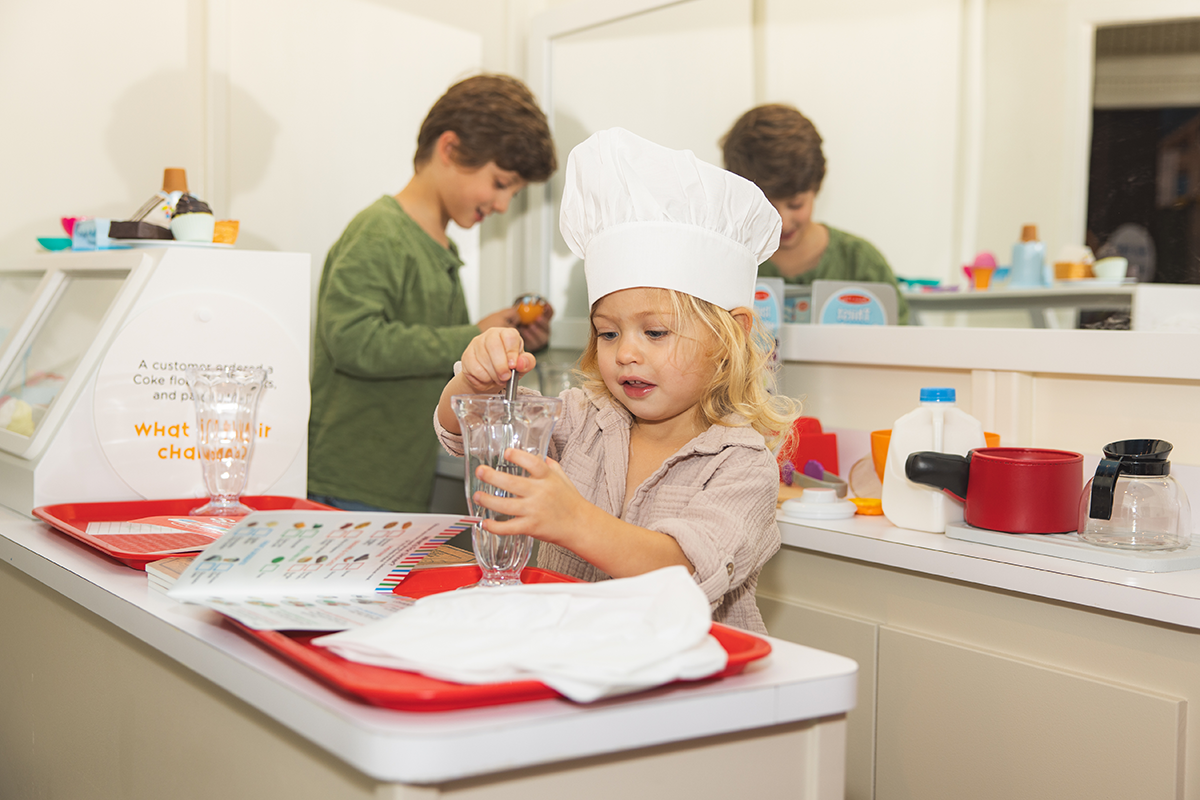 Photo: Toddler plays with milkshake glass behind a diner counter while another slightly older child plays in the kitchen behind her.