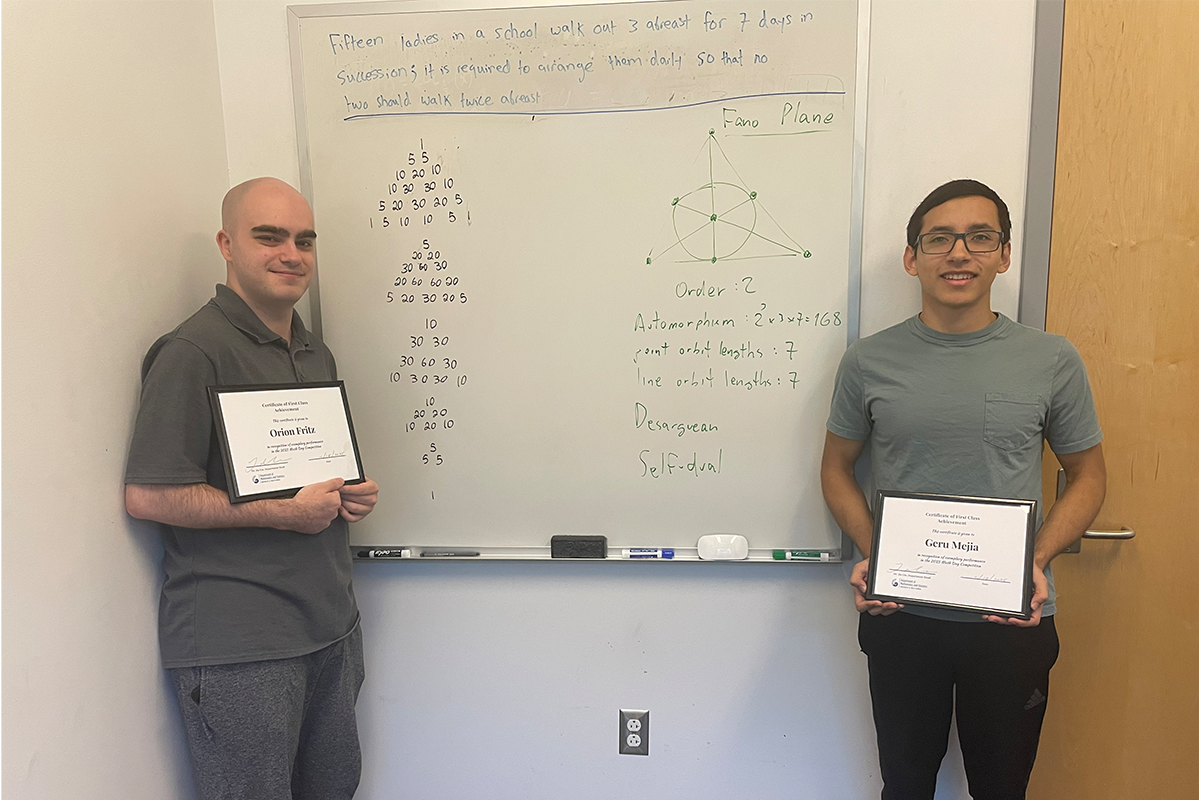 Photo: Two students stand on each side of a whiteboard with a math problem.
