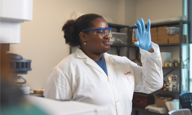 Student in white lab coat and safety goggles looks at vial in science lab.