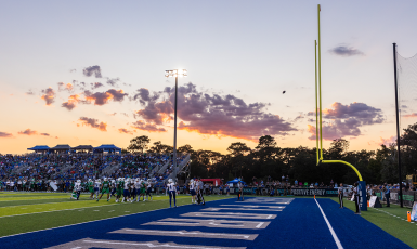 A football flies towards the goalposts on PenAir field while players and spectators look on and the sun sets.