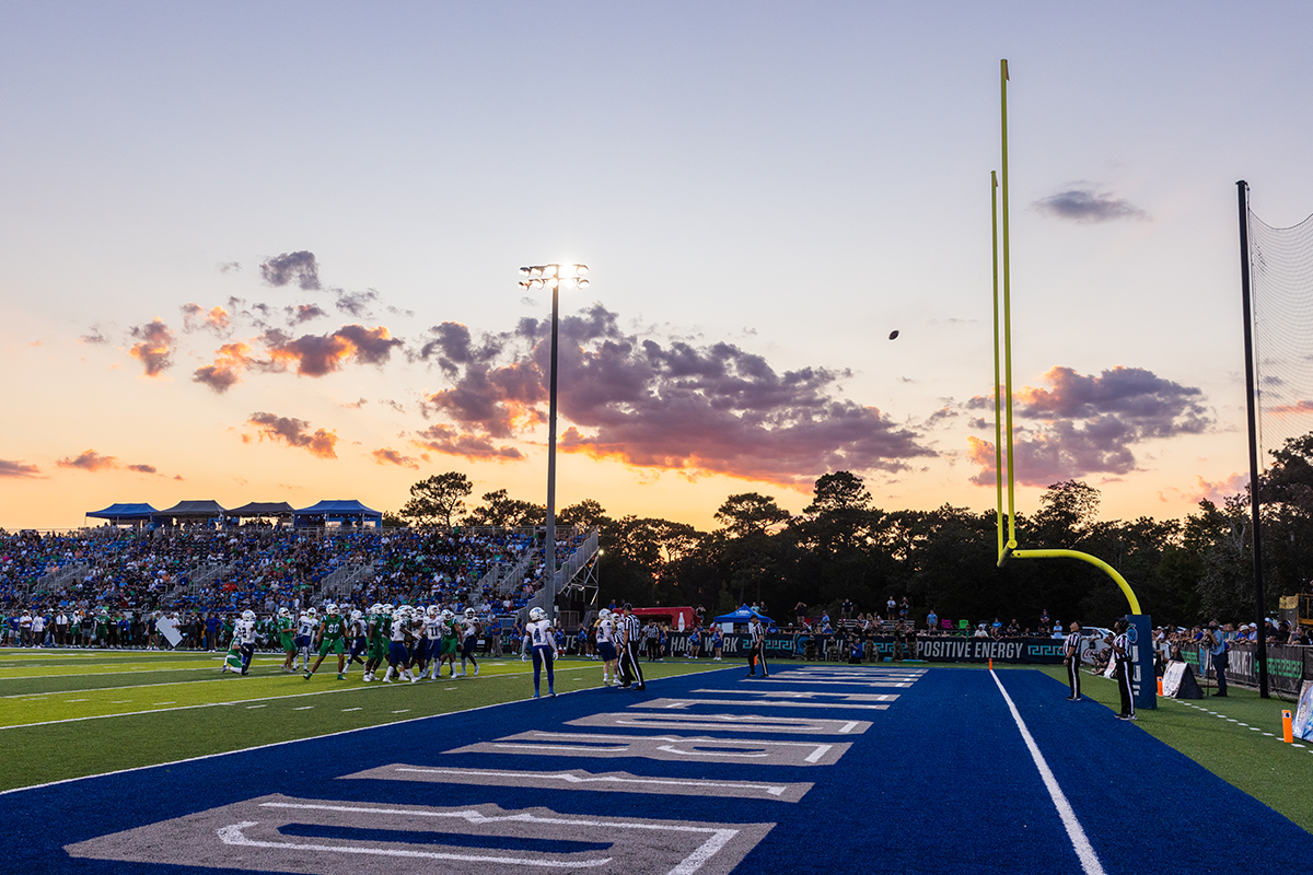 A football flies towards the goalposts on PenAir field while players and spectators look on and the sun sets.